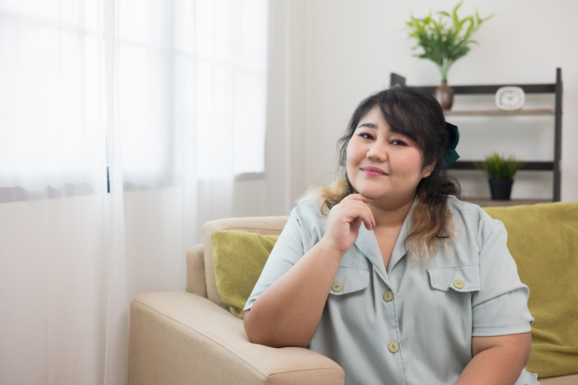 Woman in a light blue shirt resting her chin on her hand while sitting on a sofa at home