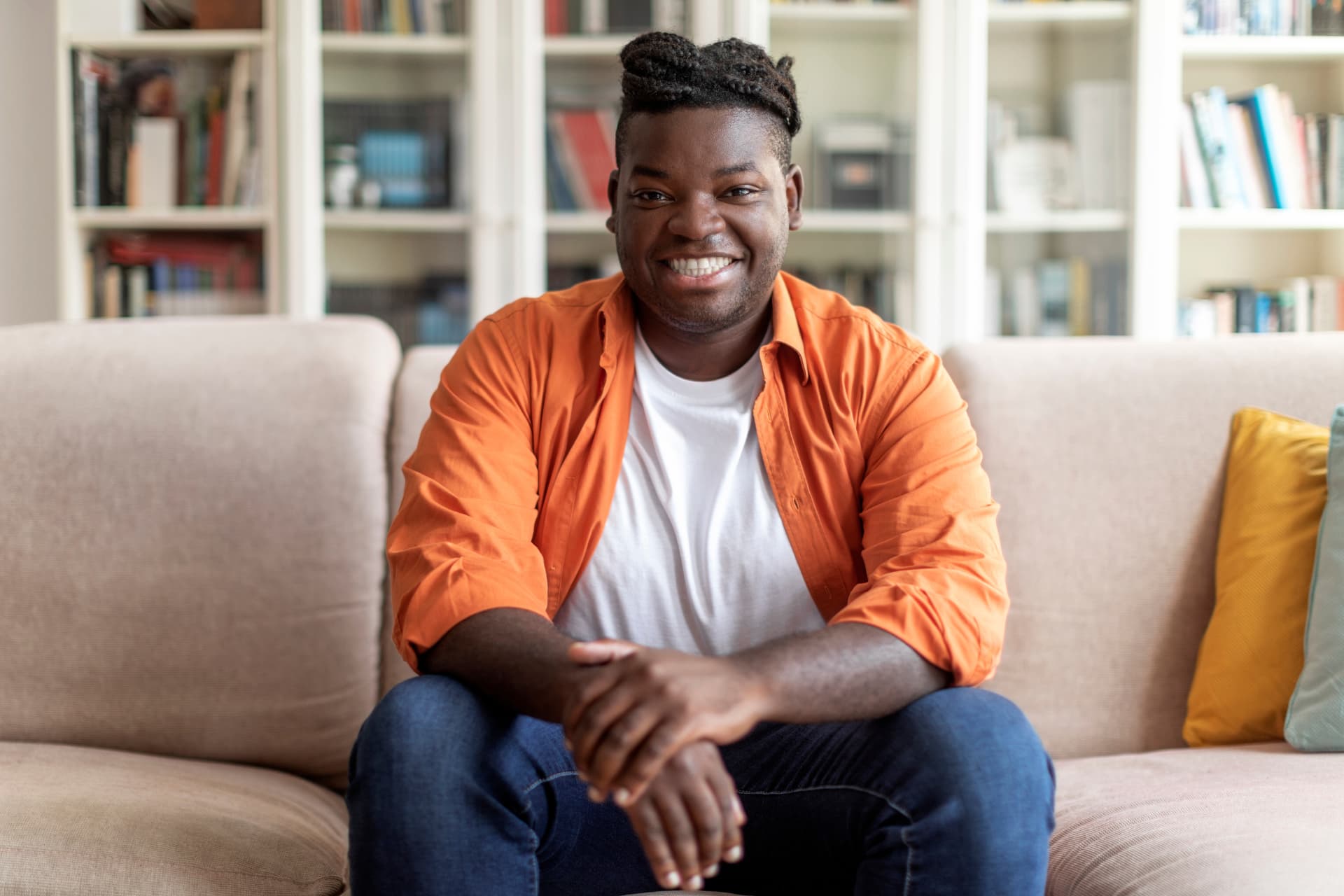 Smiling man in an orange shirt sitting on a couch in front of bookshelves