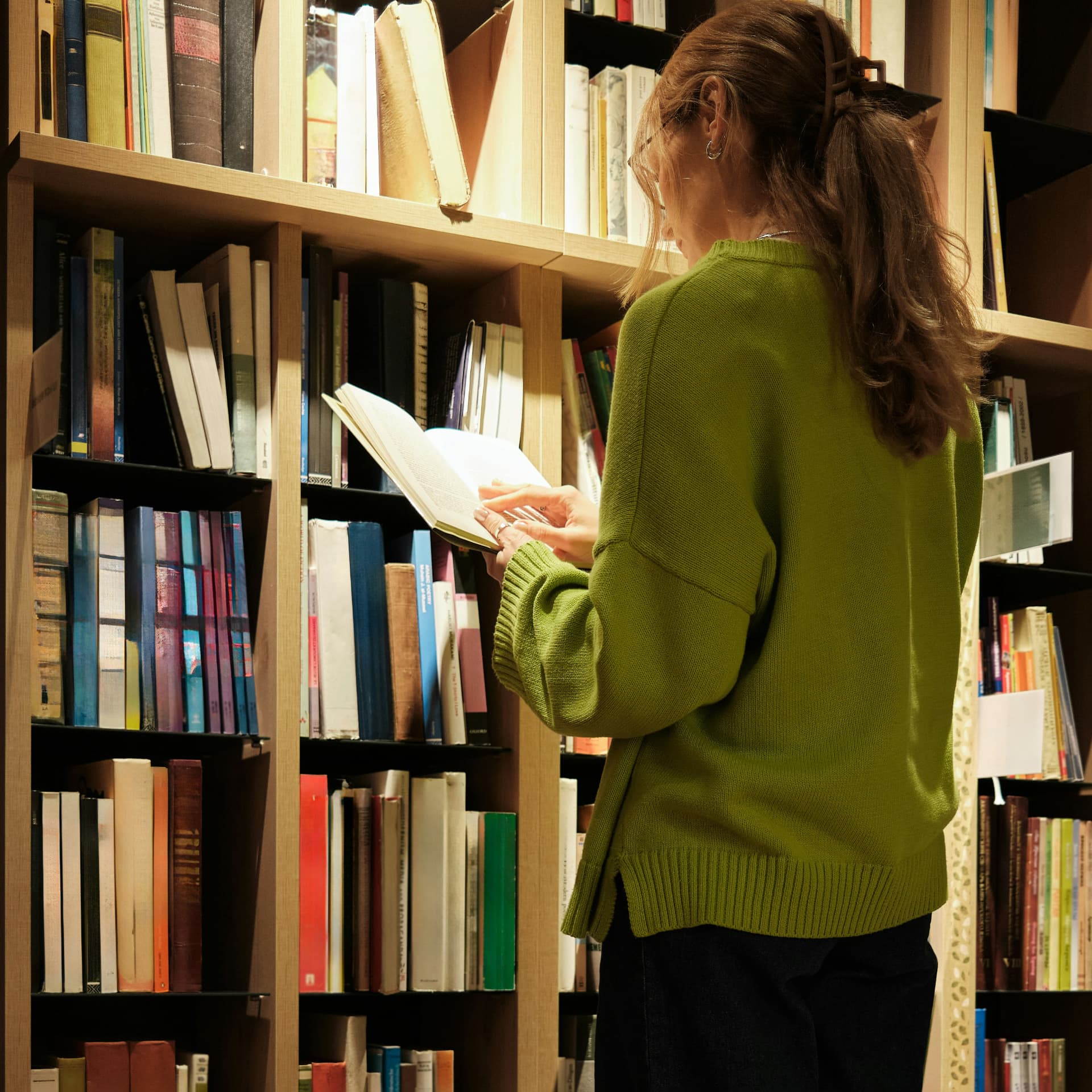 Woman in a green sweater reading an open book in front of tall bookshelves