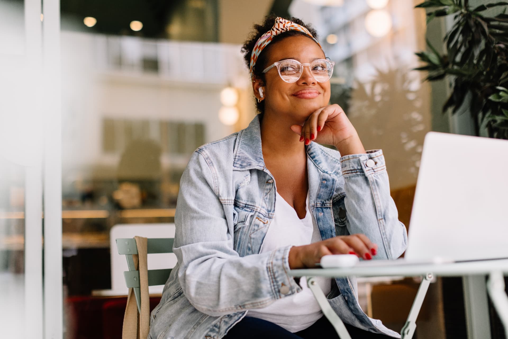 Smiling woman in a denim jacket and glasses working on a laptop at a cafe table