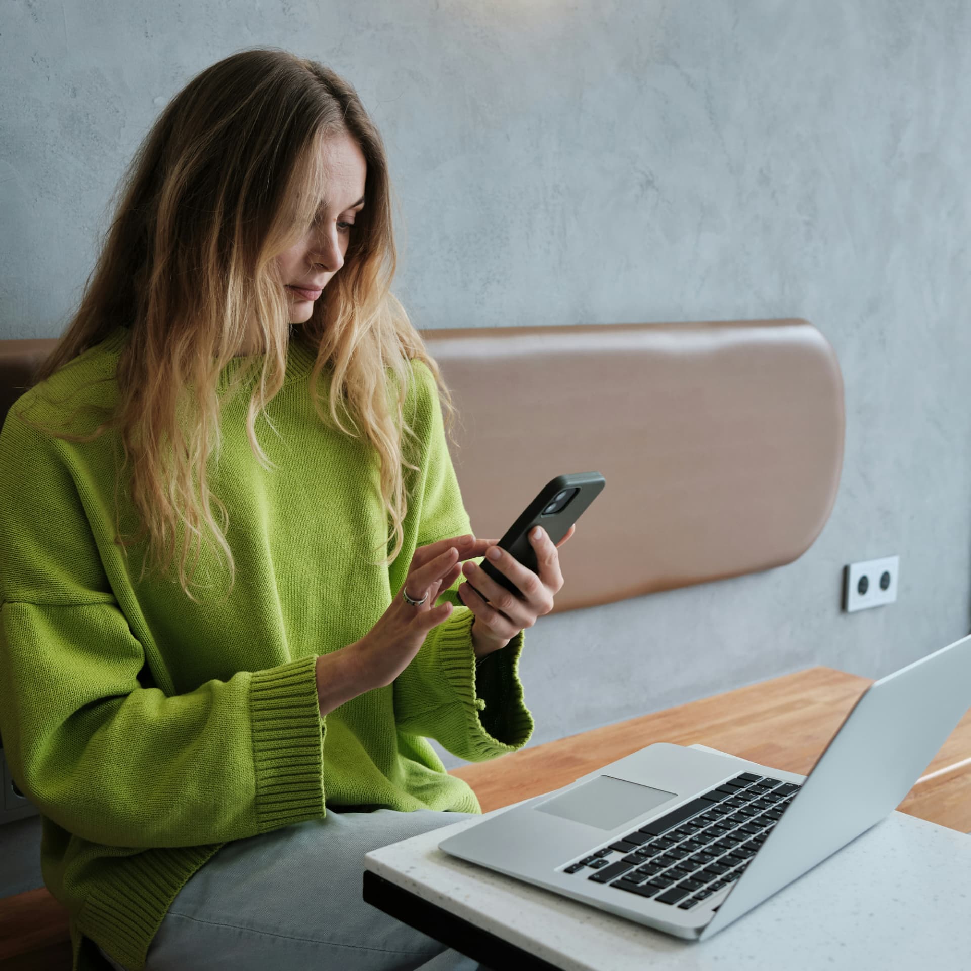 Woman in a green sweater checking her phone beside a laptop at a cafe table