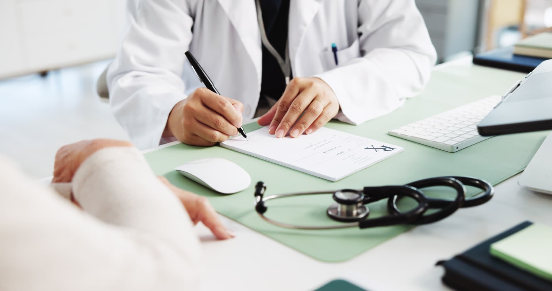 Clinician in a white coat writing a prescription at a desk beside a stethoscope