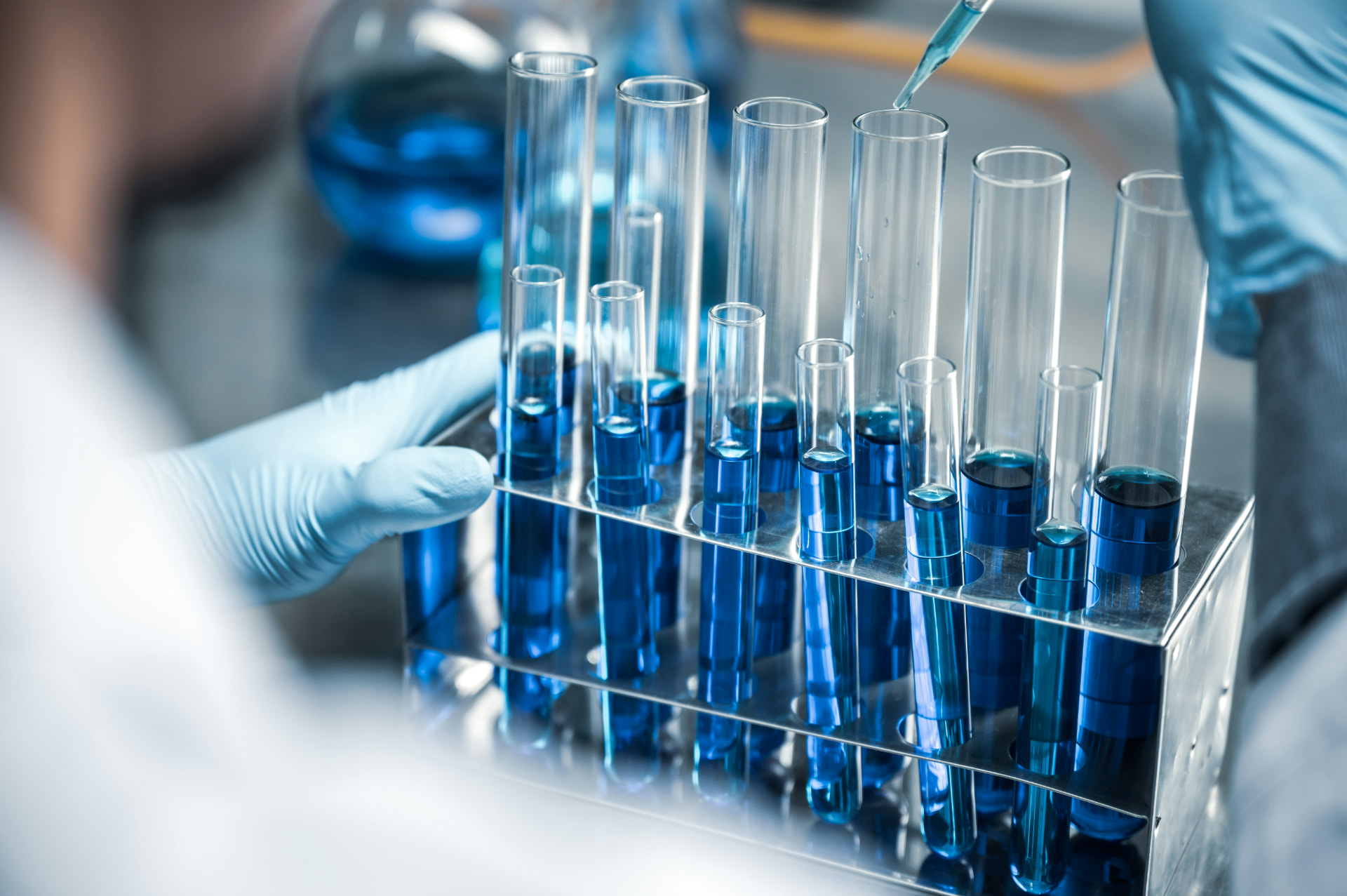 Gloved lab scientist adding liquid by pipette to a rack of test tubes filled with blue solution